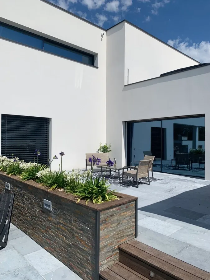 Terrasse d’une maison contemporaine blanche avec baie vitrée, volet noir et jardinière en pierre