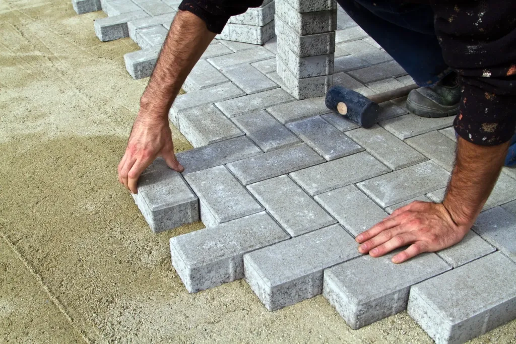 Mains posant des pavés gris en chevrons sur un lit de sable, avec un maillet