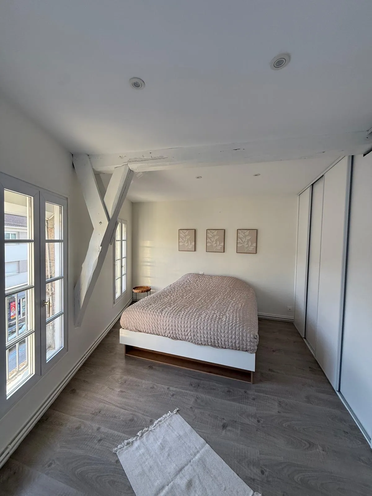 Chambre aux murs blancs avec poutres blanches apparentes, lit double et parquet gris