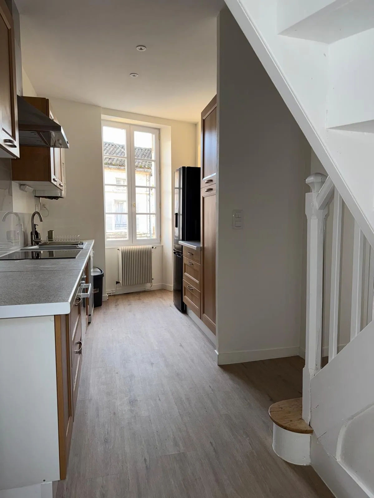 Cuisine en longueur aux meubles en bois, fenêtre au fond et escalier blanc à droite
