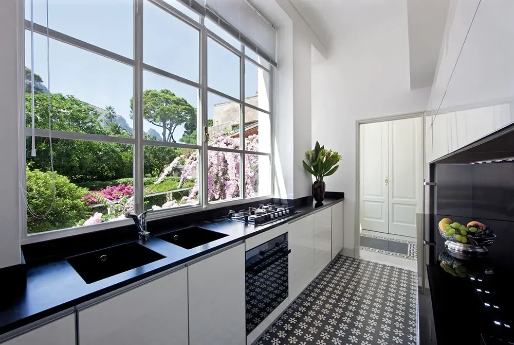 Cuisine étroite aux meubles blancs et plan de travail noir, grande fenêtre sur un jardin fleuri et sol en carreaux de ciment