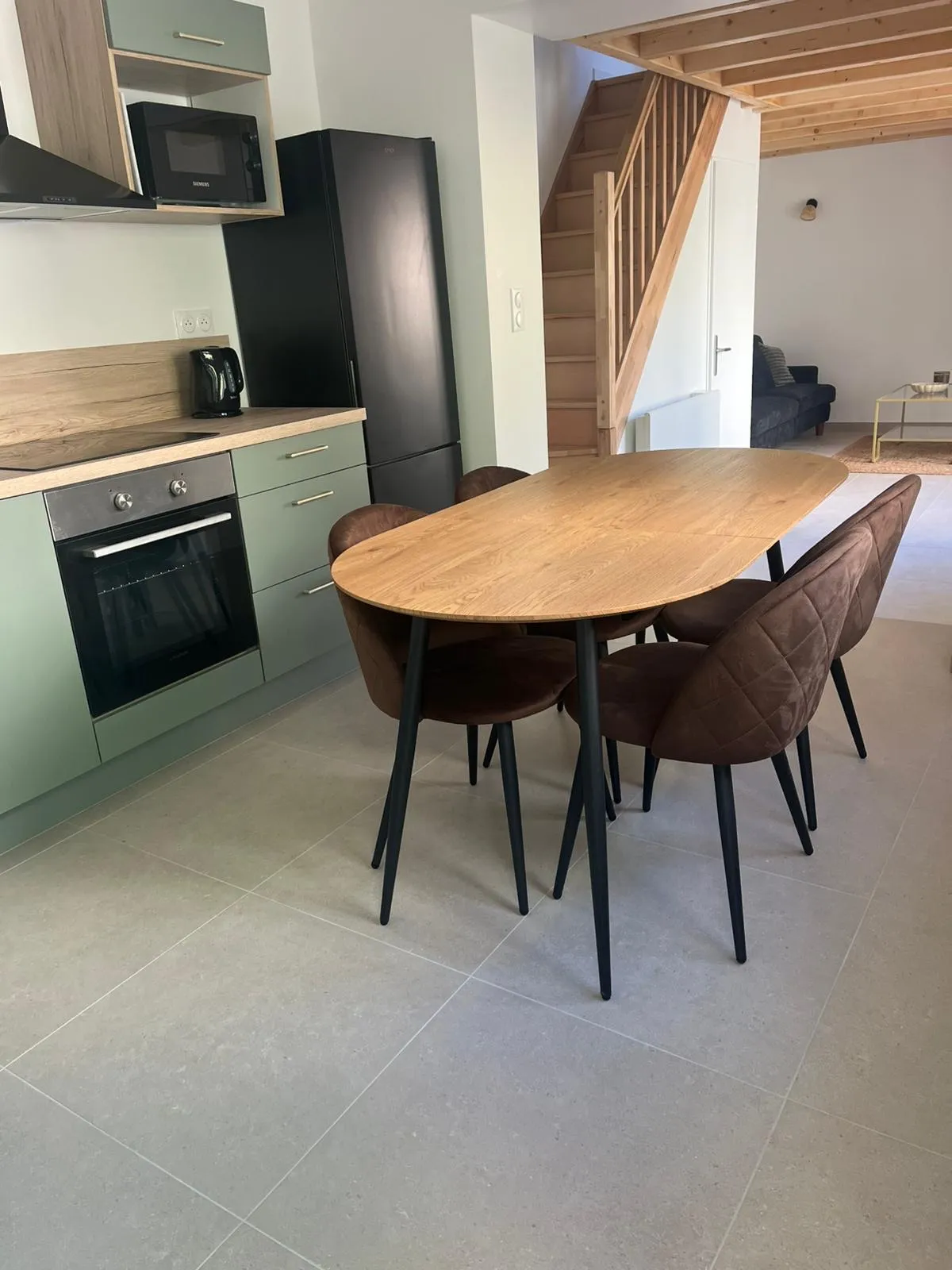 Cuisine vert sauge ouverte sur une table ovale en bois et quatre chaises, sol carrelé gris, escalier en bois au fond