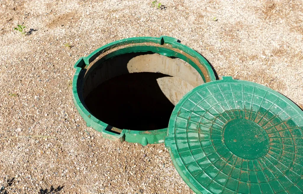 Regard de fosse septique ouvert, couvercle vert posé à côté, sur un sol de gravier