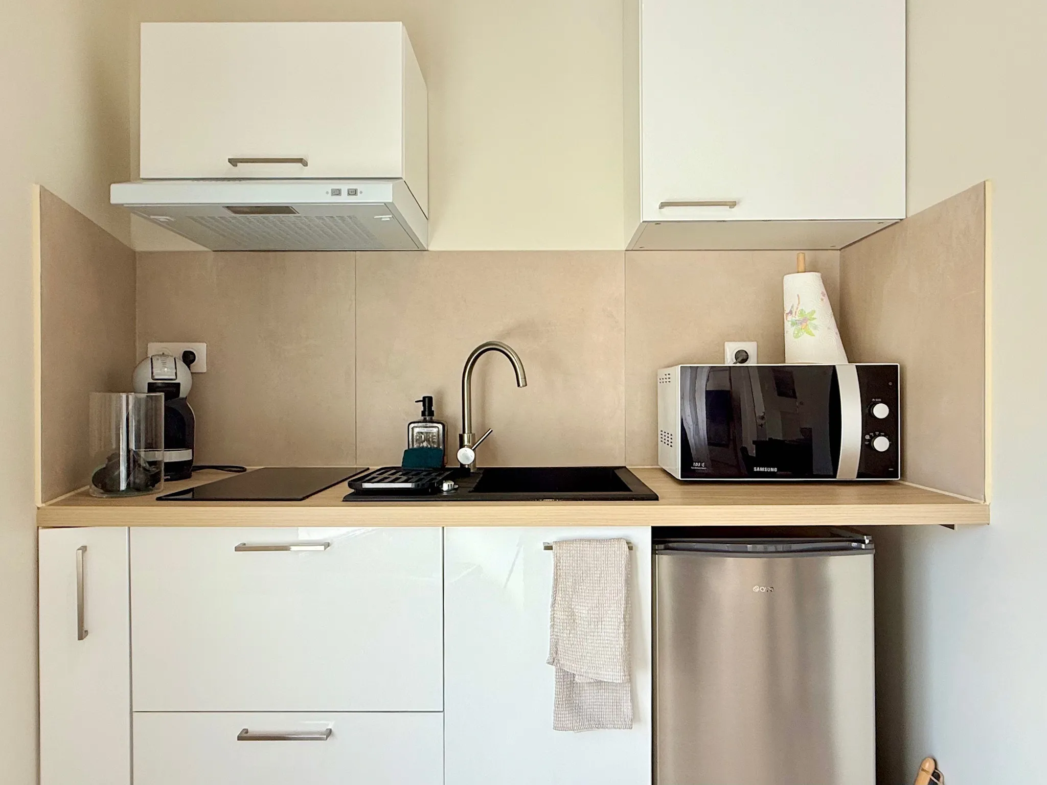 Kitchenette blanche avec crédence en grands carreaux beiges, plaque, évier noir et réfrigérateur table top
