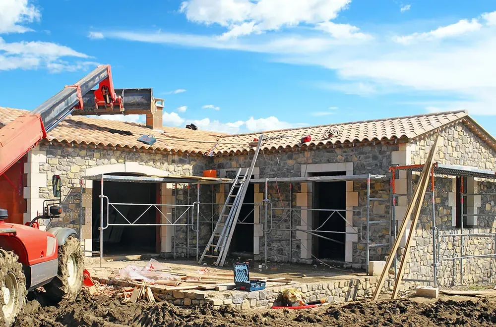 Maison en pierre couverte de tuiles pendant le chantier, avec échafaudage et chariot élévateur sous un ciel bleu