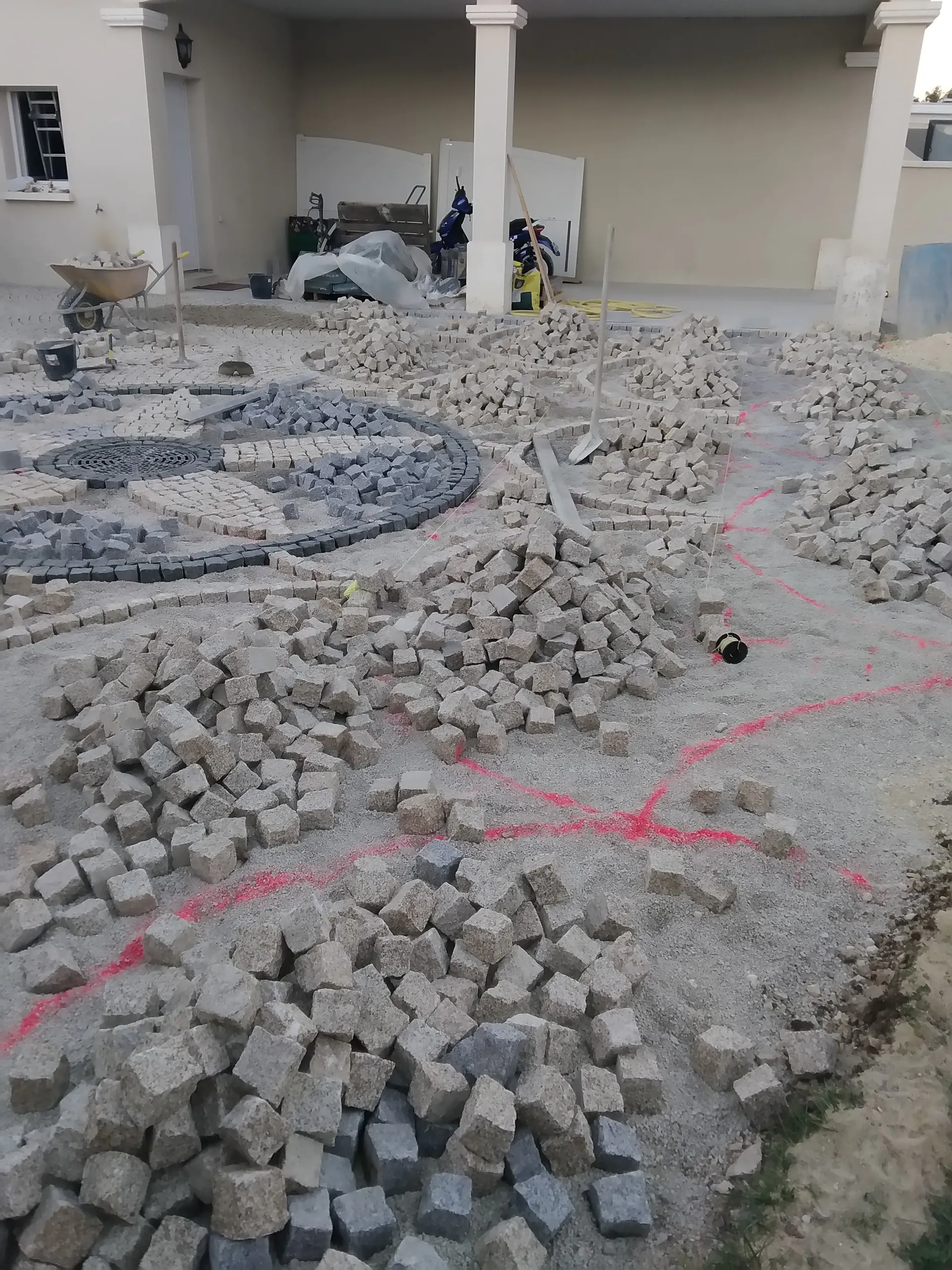 Pavés en granit en tas et premiers rangs posés sur le sable, avec des repères de traçage rouges au sol