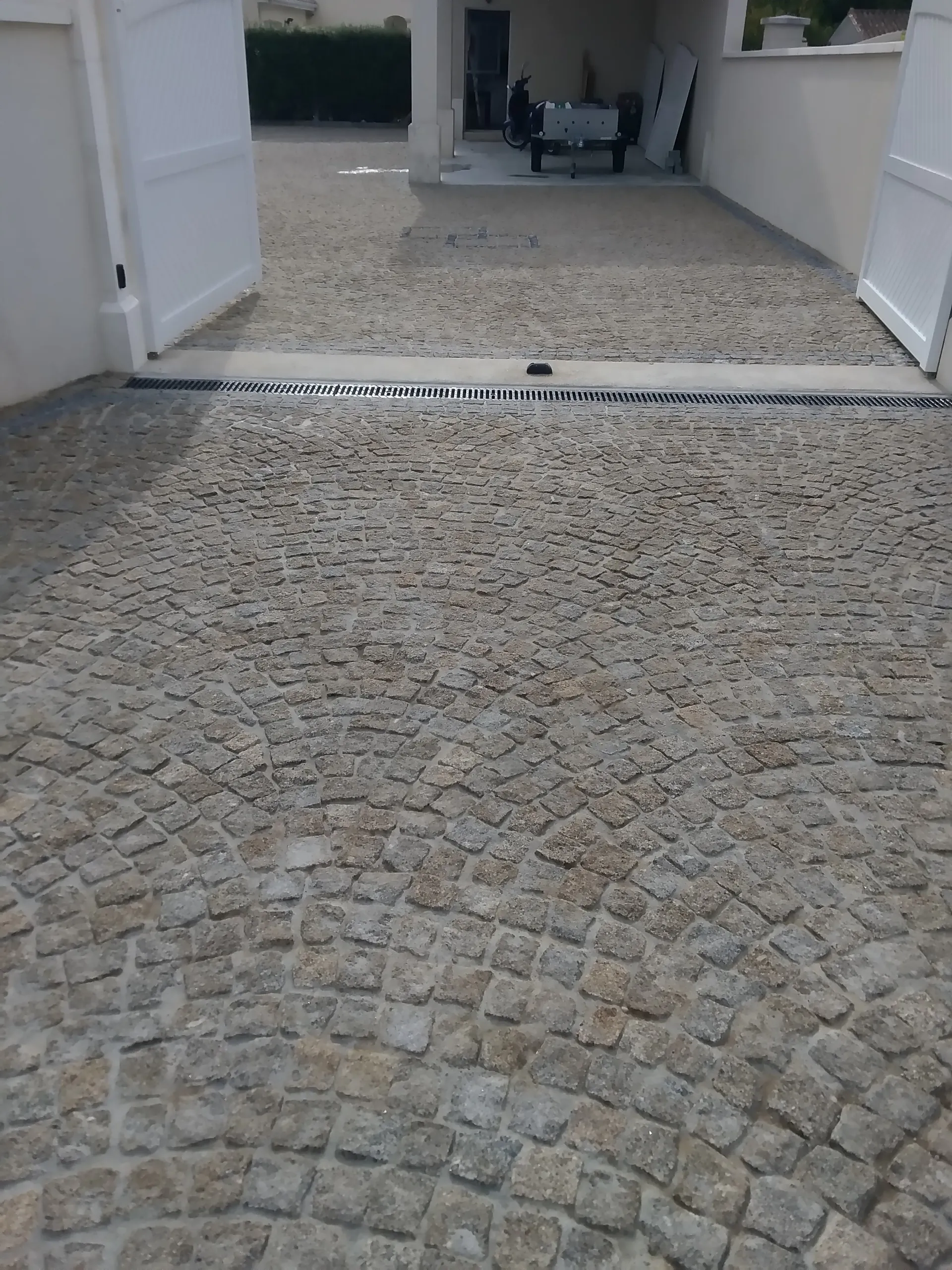 Cour pavée en granit terminée, avec un caniveau à grille devant un portail blanc ouvert