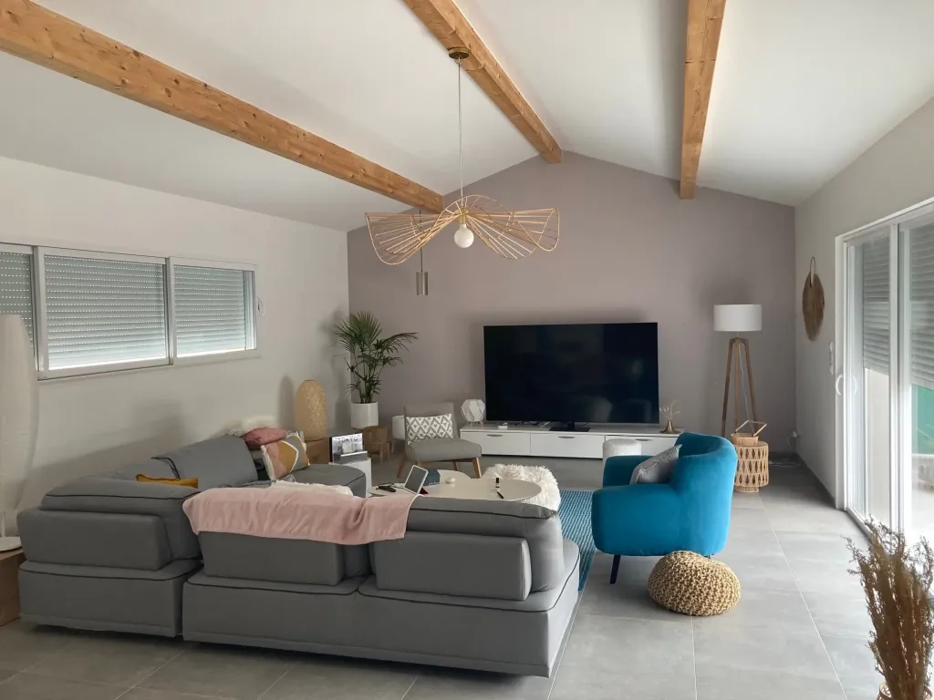 Salon sous charpente apparente aux poutres en bois clair, canapé gris, fauteuil bleu et baie coulissante
