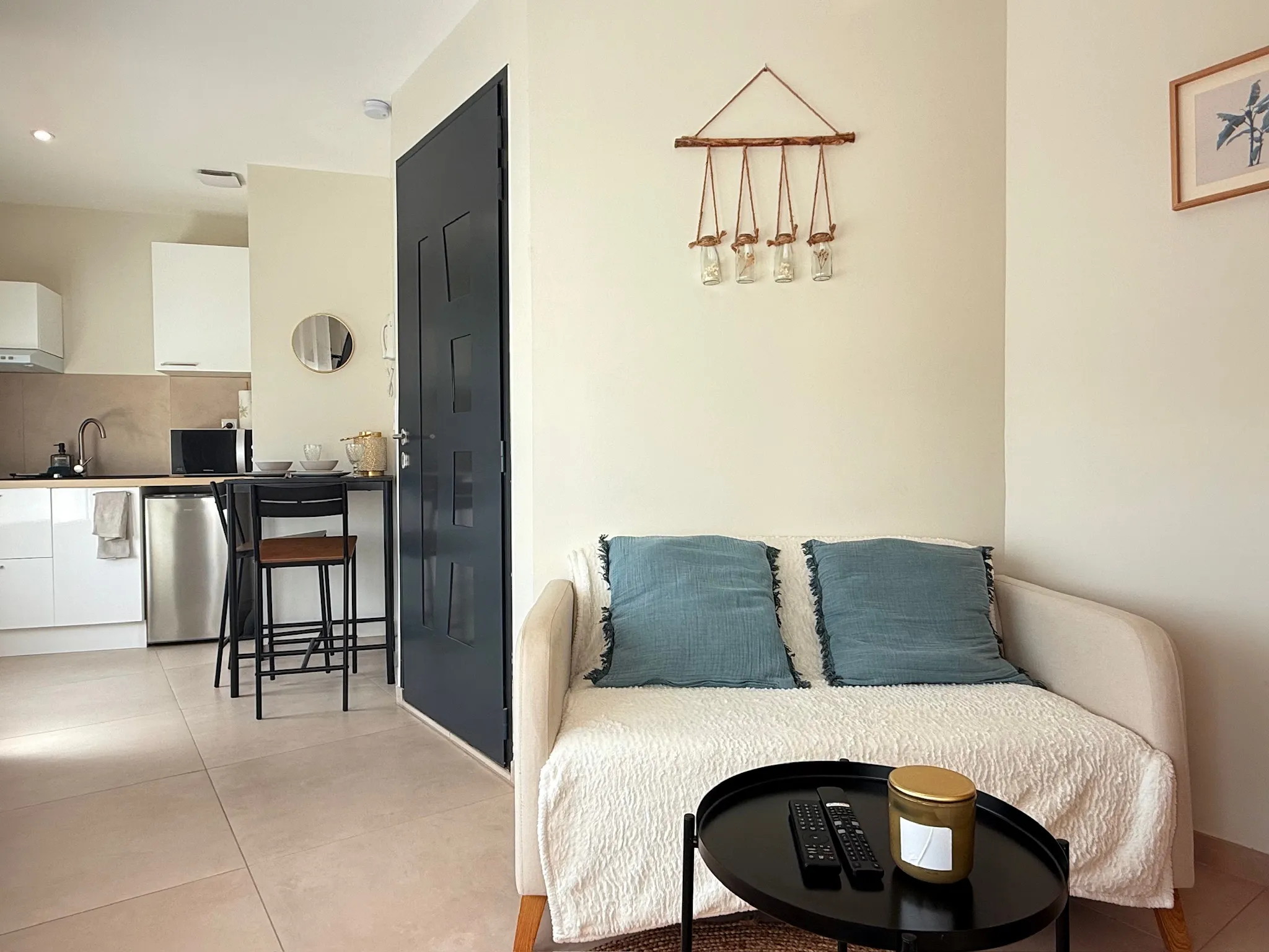 Studio lumineux avec canapé beige et coussins bleus, coin cuisine blanc et porte d'entrée gris anthracite