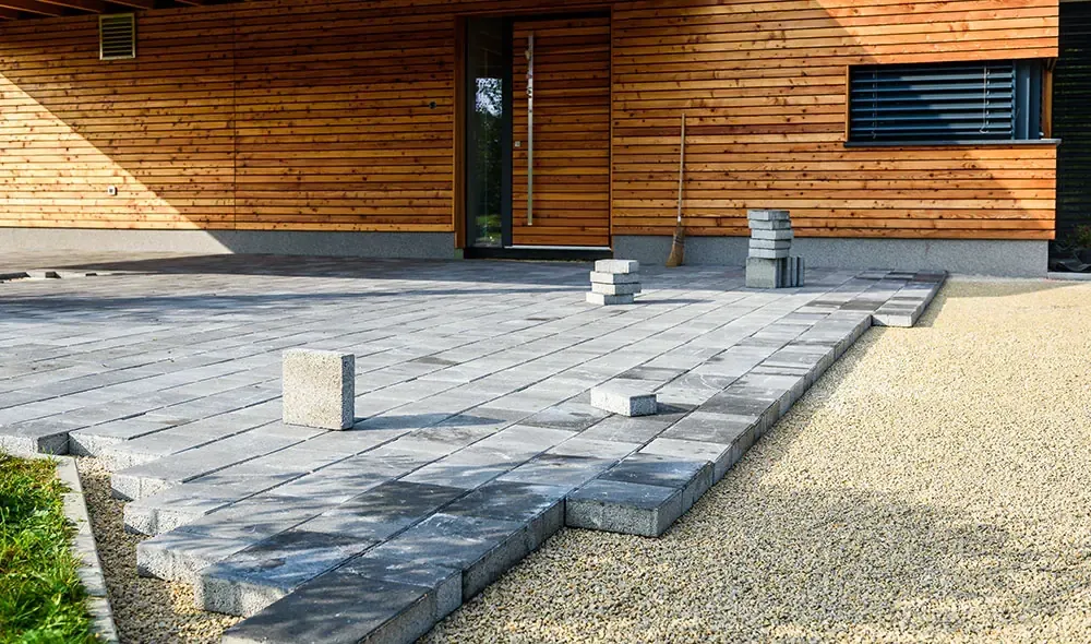Terrasse en cours de pose de dalles grises, avec bordure de gravier et façade en bardage bois