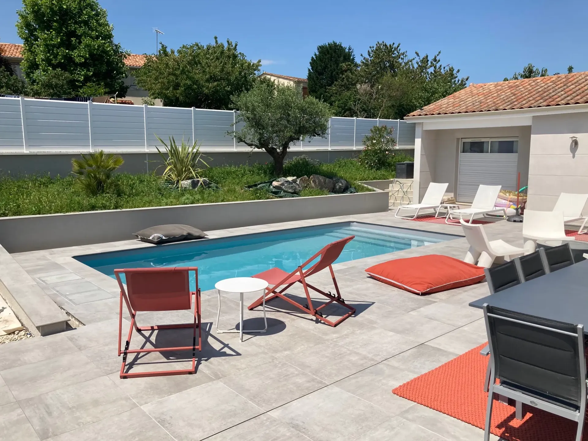Piscine rectangulaire entourée de dalles gris clair, avec chaises longues, fauteuils rouges et muret végétalisé