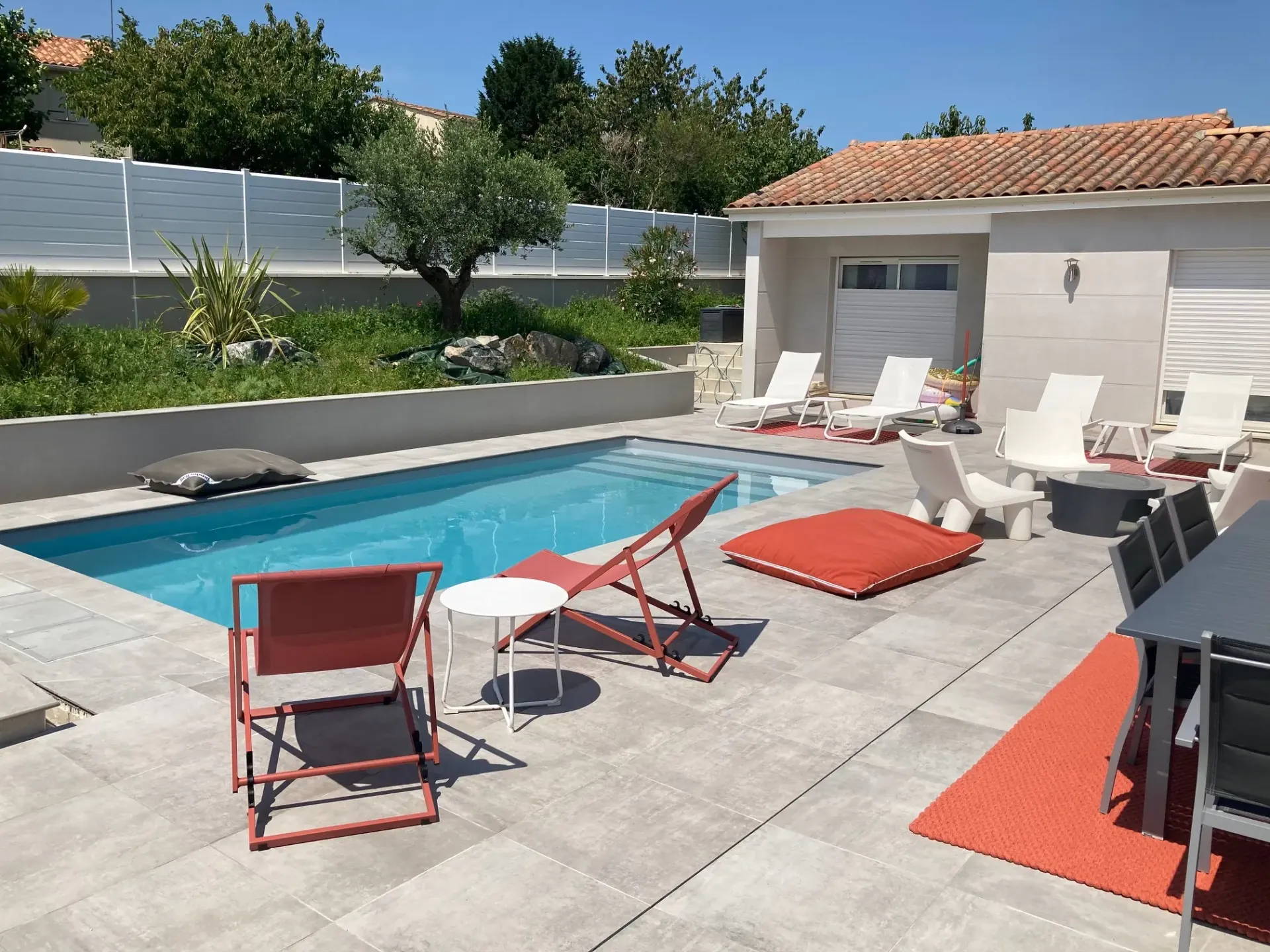 Tour de piscine en dalles gris clair vu de près, avec transats et coussin orange devant une dépendance