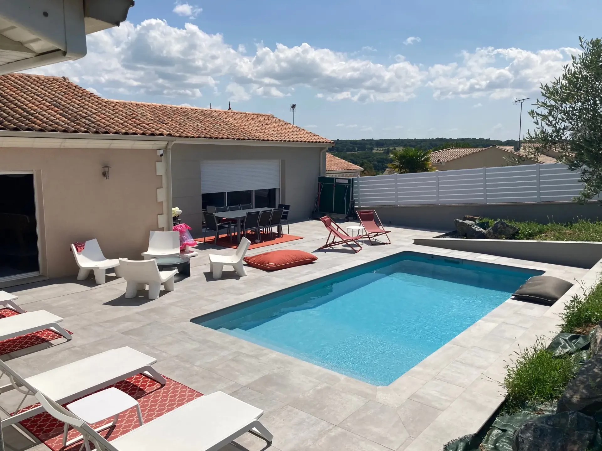 Terrasse en dalles gris clair autour d'une piscine, devant une maison à toit de tuiles