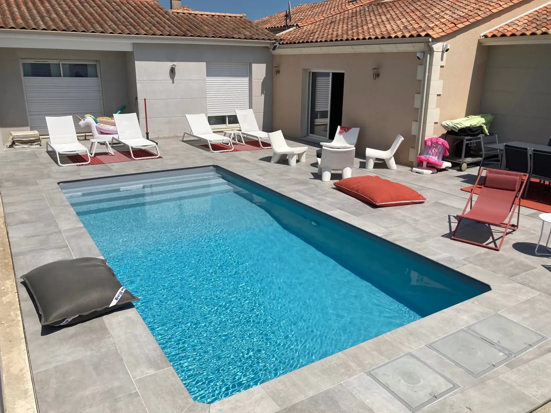 Piscine entourée de dalles gris clair dans une cour fermée, avec chaises longues et mobilier de jardin