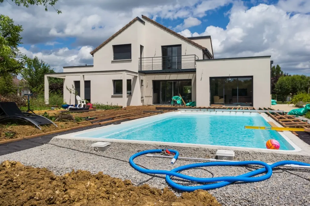 Maison avec piscine bleue et terrain encore en terre battue, tuyau de nettoyage au premier plan